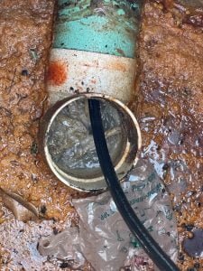 Sewer pipe with a clog being cleared by a black hose, surrounded by muddy ground.